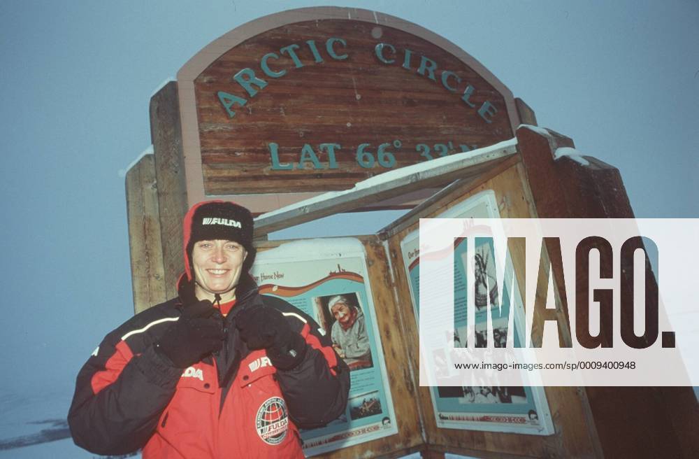Jutta Kleinschmidt am Polar- kreis im Yukon-Territorium (Kanada) 02 00 mapo Frau Motorsportlerin Motorsport Auto Autosport Rallyes Rallye Rallyesport Rallyefahrerin Rallyefahrer Fahrerin Fahrer blond leger sportlich Jacke Anorak rot-schwarz Logo Fulda Mütze Handschuhe stehend draussen Schnee halb quer lächelnd posierend im Hintergrund Schild Arctic Circle Polarkreis