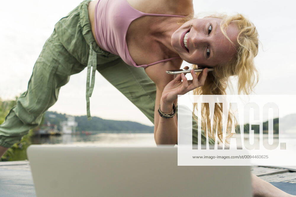 Young woman stretching and using laptop and cell phone on a jetty at a lake model released Symbolfoto JOSF03614
