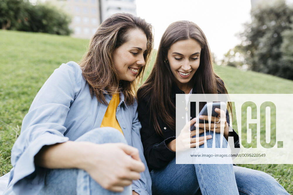 Two friends looking on the smartphone, sitting on a meadow of a park model released Symbolfoto JRFF03803