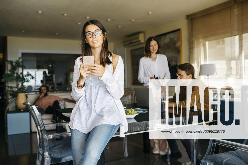 Portrait of woman with cell phone and friends working in background at home model released Symbolfoto property released JRFF03890
