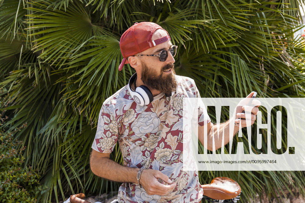 Mature man with red basecap, sunglasses and white headphones using smartphone model released Symbolfoto TCF06224