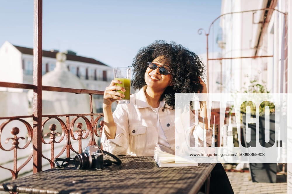 Smiling young woman with curly hair sitting at table on balcony with smoothie and camera model released Symbolfoto property released DCRF00104