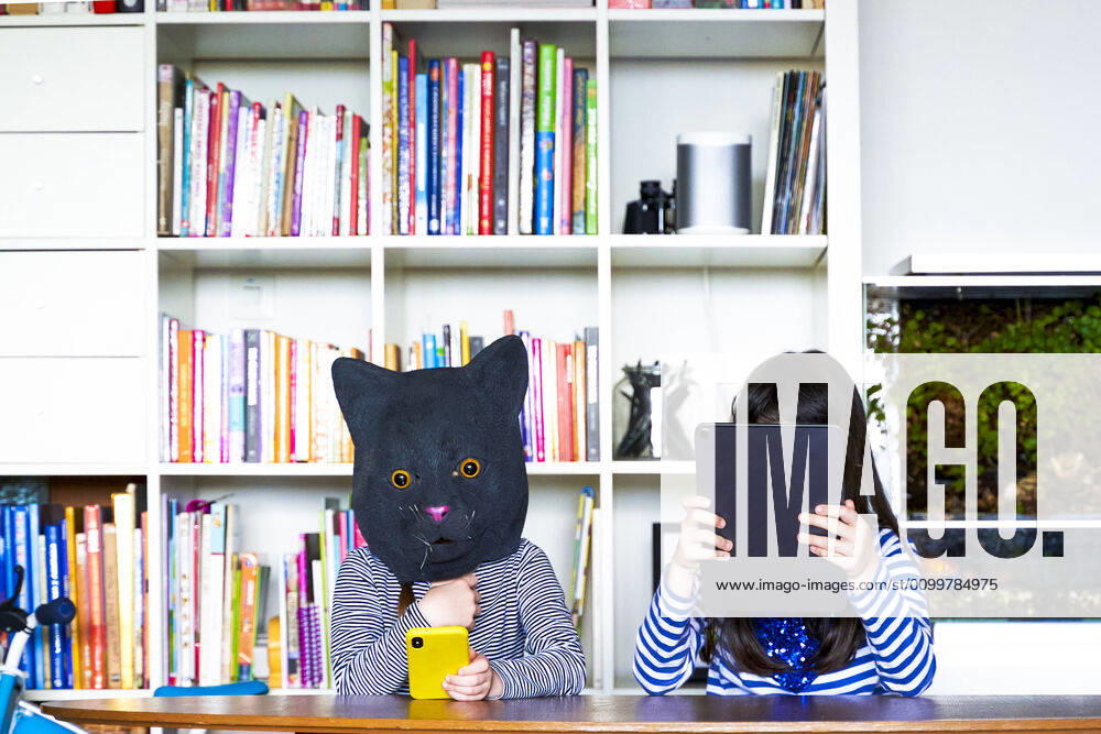 Two girls sitting at table, hiding behind digital tablet and cat mask model released Symbolfoto property released ERRF03439