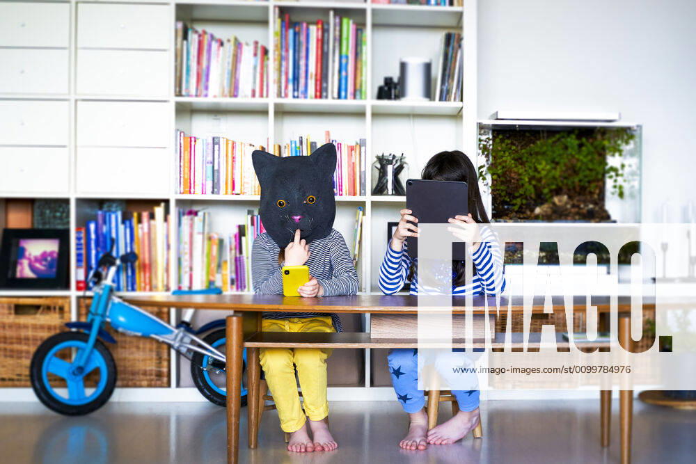 Two girls sitting at table, hiding behind digital tablet and cat mask model released Symbolfoto property released ERRF03440