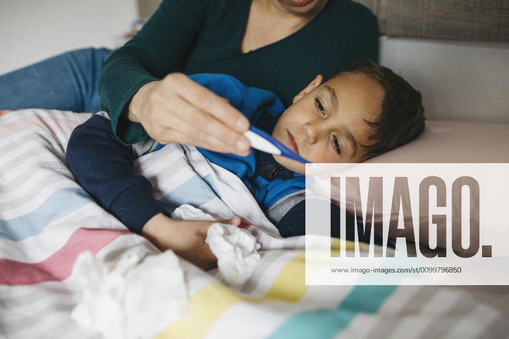 Sick boy with tissue in his hand lying in bed looking together with his mother at digital thermometer model released Symbolfoto property released JRFF04244