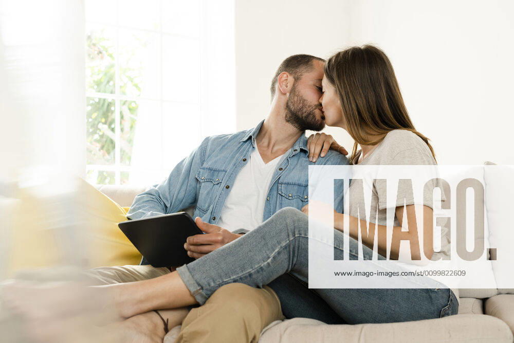 Affectionate couple relaxing at home and kissing while sitting on couch and holding tablet model released Symbolfoto property released SBOF02192