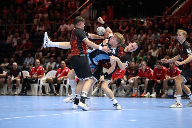 An HSV Hamburg player in a light blue and dark blue jersey drives past two MTV Braunschweig defenders with the ball in his hand. The matchup takes place in front of a packed crowd at the Volkswagen Halle in Braunschweig. The HSV Hamburg player fights his way through the MTV Braunschweig defense. Copyright: xspeedshotx