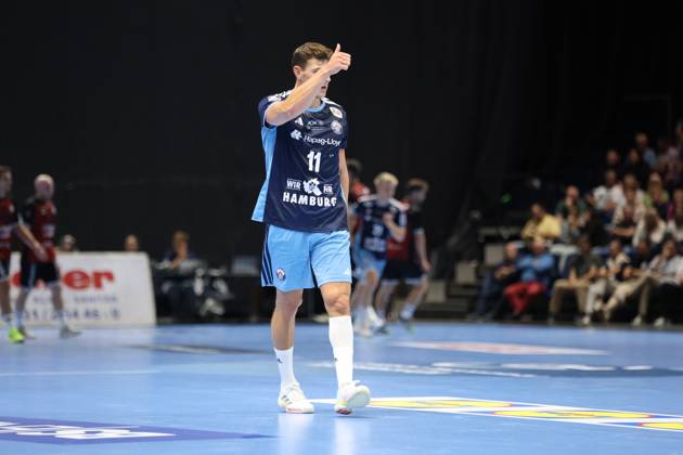 Nicolaj Jørgensen (No. 11, HSV Hamburg) stands on the court at the Volkswagen Halle in Braunschweig and gives a thumbs-up, raising his hand to his face. In the background, other players from both teams and spectators in the stands can be seen. Nicolaj Jørgensen (No. 11, HSV Hamburg) gives a thumbs-up. Copyright: xspeedshotx