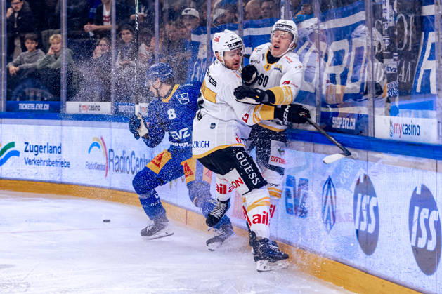 Rico Gredig 8 (EV Zug) evades a check by Mirco Müller 25 (HC Lugano); instead, Lassi Thomson 60 (HC Lugano) is hit. 27 10 2026 National League, Regular Season, Ice Hockey, OYM Hall, EV Zug vs. HC Lugano. Photo by  Copyright: xJustPictures.ch PhilippxHegglinx