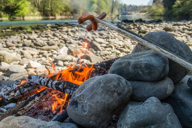 Bei einer Fahrt mit dem Kajak im Frühling entlang der Iller macht Lilli eine Pause und grillt am Lagerfeuer eine Wurst. Altusried, Bayern, Deutschland. Frühlingsfahrt auf einem Fluss mit dem Kajak.