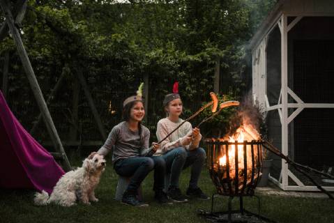 Two girls wearing feather headdress, grilling sausage over camp fire, dog sitting on meadow model released Symbolfoto property released  MOEF01216
