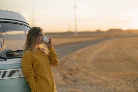 Young woman drinking from mug at camper van in rural landscape at sunset model released Symbolfoto  GUSF01634