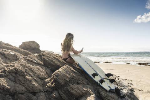 Young woman with surfboard sitting on a rock at the beach, using smartphone model released Symbolfoto  UUF16485
