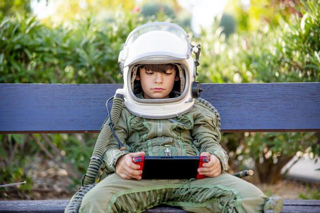 IMAGO Creative, Lifestyle, Gaming Creative Highlights Symbolfotos Boy wearing a space suit and sitting on a bench, playing alone with video games model released Symbolfoto  CJMF00124