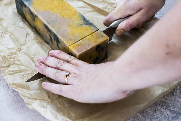 High angle close up person cutting yellow and black homemade bar of soap with kitchen knife. High angle close up person cutting yellow and black homemade bar of soap with kitchen knife.  59315