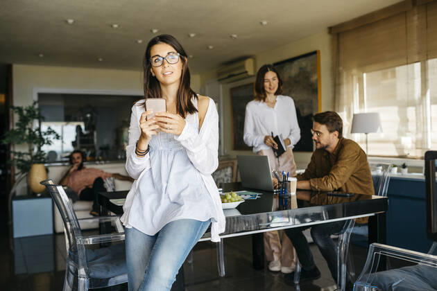 Portrait of woman with cell phone and friends working in background at home model released Symbolfoto property released  JRFF03890