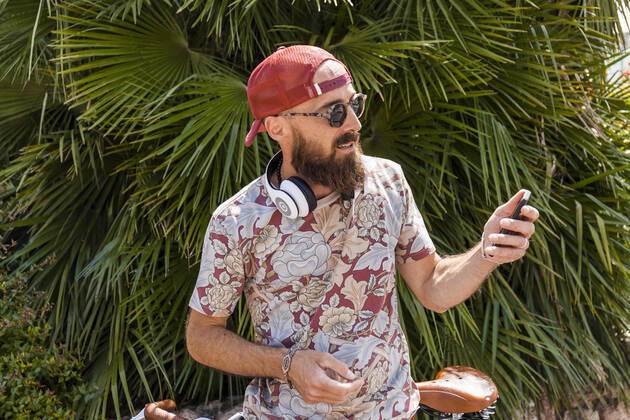 Mature man with red basecap, sunglasses and white headphones using smartphone model released Symbolfoto TCF06224