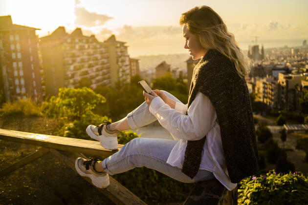 Young woman sitting on railing above the city using cell phone, Barcelona, Spain model released Symbolfoto GIOF07950