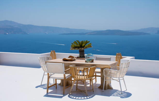 furniture composed by a table and some chairs on a white terrace of a house in Santorini Greece where you can enjoy meal while seeing a romantic seascape to the blue Aegean sea. Horizontal ph Oia, Greece CR_RUBO200219-310702-01
