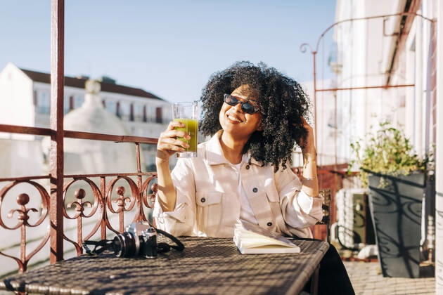 Smiling young woman with curly hair sitting at table on balcony with smoothie and camera model released Symbolfoto property released  DCRF00104