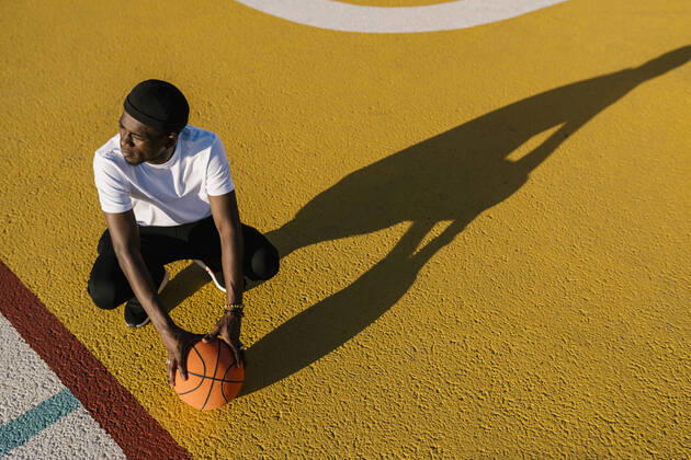 Thoughtful young man holding basketball crouching on sports court during sunny day model released Symbolfoto EGAF00294