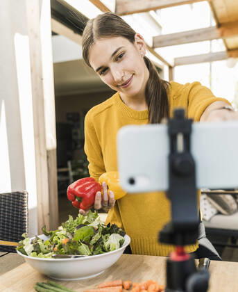 Smiling young woman adjusting smart phone on tripod while filming at home model released Symbolfoto property released UUF21046