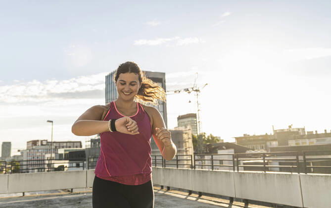 Smiling young woman checking time while running on terrace against sky model released Symbolfoto UUF21378