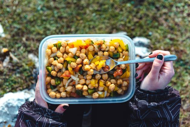 From above crop woman in warm jacket holding plastic container of chickpea with veggies having lunch on meadow while hiking in Spain, Model released Copyright: xAlbertoxMenendezx MR