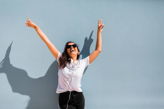 Carefree beautiful woman in casual outfit and headphones on neck standing with hands up beside blue wall of building on city street, Model released Copyright: xAlexandraxCoelhox MR
