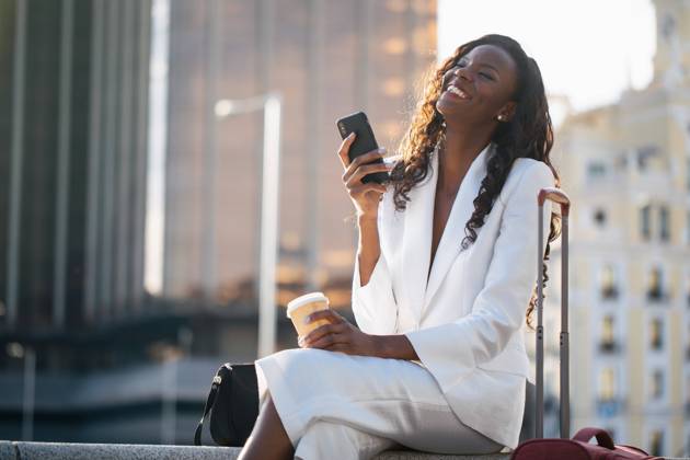 Positive African American businesswoman in elegant white clothes browsing cellphone and having coffee while sitting on street with suitcase, Model released, Property released Copyright: xFernandoxValenciax MR