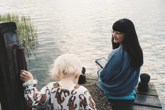 Mother looking at daughter while holding digital tablet and sitting on jetty over lake , model released, property released Copyright: xMaskotx , MA58869