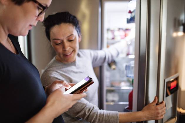 Woman showing mobile phone while girlfriend opening refrigerator in kitchen , model released, property released Copyright: xMaskotx , MA63056