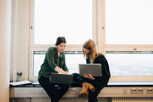 Confident female colleagues discussing over laptops while sitting on window sill at creative office , model released, property released Copyright: xMaskotx , MA63814