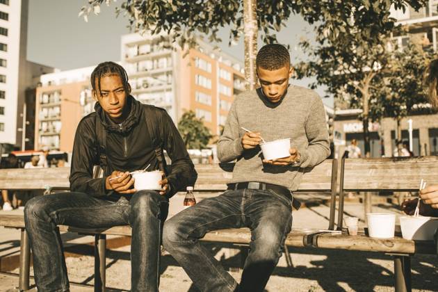 Portrait of serious teenage boy holding take out food while sitting by friend on bench in city during sunny day , model released Copyright: xMaskotx , MA69393