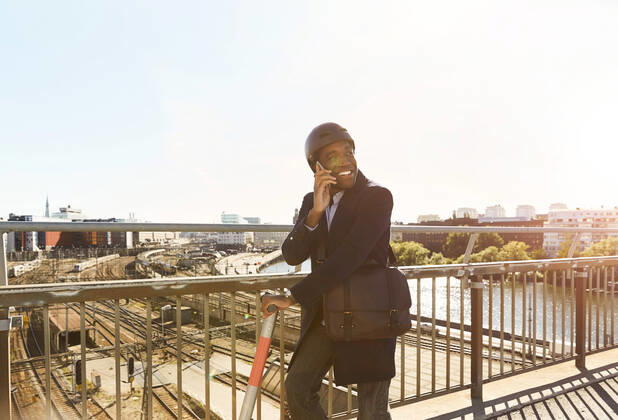 IMAGO Creative: Business, Innovative Technologie-Lösungen Smiling young man talking on smart phone while standing with electric push scooter at bridge in city against sky , model released Copyright: xMaskotx , MA69560