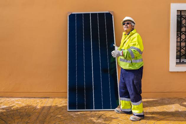IMAGO Creative: Business, Innovative Technologie-L?sungen Side view of mature experienced male technician in uniform and helmet standing with solar panel near yellow building while working on installation of renewable energy system Copyright: xJavierxDexLaxTorrex Model Released