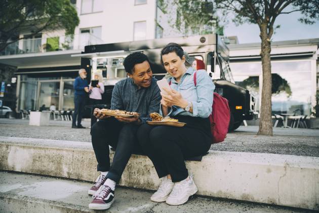 Smiling friends sharing smart phone while sitting with meal against food truck in city Sweden, Stockholm, model released, property released Copyright: xMaskotx , MA81566