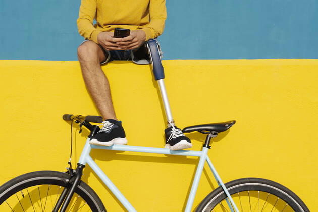 IMAGO Creative: Gesundheit, Prothese Young disabled man with bicycle using mobile phone while sitting on multi colored wall model released Symbolfoto JCZF00287