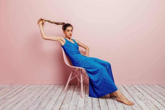 Side view of young woman in blue dress barefoot looking at camera holding ponytail while sitting with in chair and looking at camera on minimalist pink background Copyright: xMarcosxOsoriox Model Released