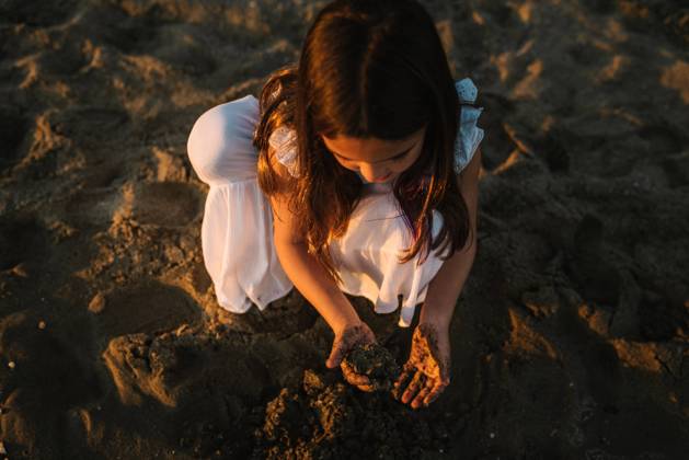 IMAGO Creative, Jahreszeit Sommer, Sommerferien Cute pensive female kid in white dress playing with sand on seaside in sunlight, Model released Copyright: xPatriciaxBecarotox MR