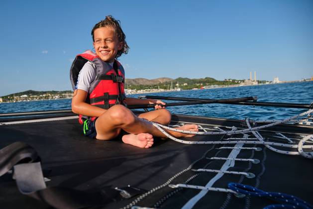 Blond kid with life jacket sitting in a boat holding the rudder and a rope while smiling and the shore far away in the background, Model released Copyright: xPhilippexDegrootex MR