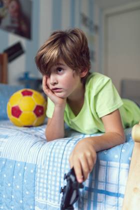 Bored little boy in casual clothes lying on bed near ball and skateboard unhappy with self isolation at home Copyright: xSergioxVictorxVegax Model Released