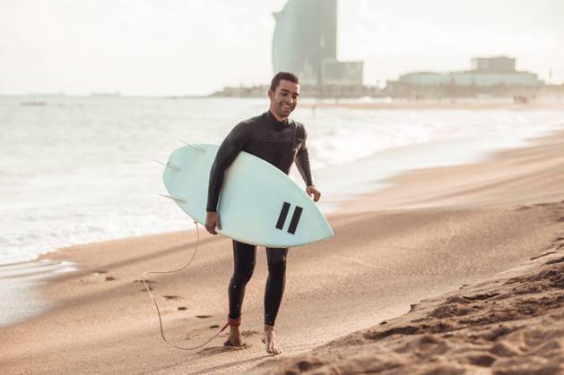 IMAGO Creative, Jahreszeit Sommer, Sommer in der Stadt Happy adult man in wetsuit carrying surfboard walking on sandy beach in sunlight, Barcelona Copyright: xAitorxLamadridx Model Released