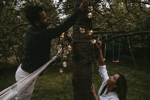 Male and female friends hanging lighting equipment in backyard during dinner party Sweden, Stockholm, model released, property released Copyright: xMaskotx , MA83390