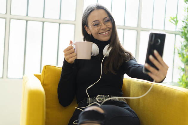 Smiling young female blogger taking selfie with coffee cup and headphones against window at home model released Symbolfoto property released GIOF09444