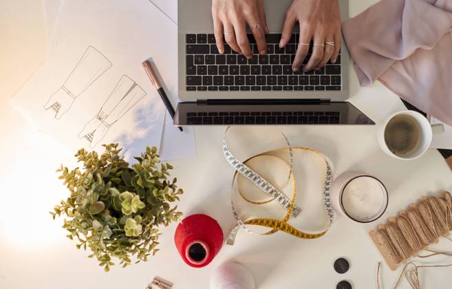 Fashion designer using laptop by fabric swatch and coffee at desk in atelier model released Symbolfoto property released VEGF03439