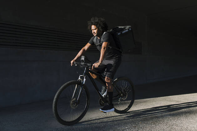 IMAGO Creative, Sommer, Schatten-Pl?tze bei Hitze Side view of ethnic male with delivery box riding bicycle on walkway against wall with shadow in sunlight Copyright: xLightsyx Model Released