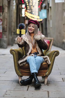 Smiling woman in fur coat vlogging through smart phone while sitting on armchair at alley model released Symbolfoto AGOF00083