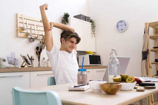 Young man stretching his hand while sitting at table with laptop in kitchen model released Symbolfoto property released GIOF11802
