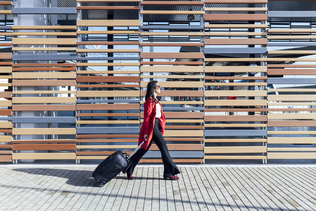 Businesswoman walking on footpath with suitcase by wooden wall during sunny day model released Symbolfoto JRVF00340